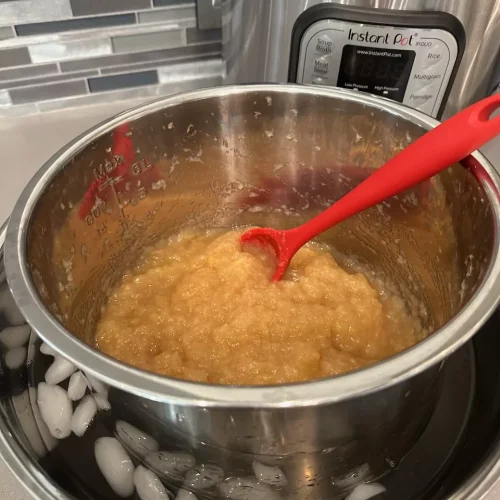 Homemade Instant Pot applesauce cooling in an ice bath with a red spoon, next to an Instant Pot on the counter. No sugar added.